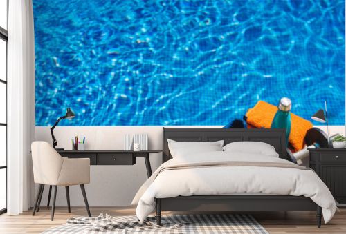 sport accessories in front of a swimming pool. orange towel, blue water bottle, weights. background of a swimming pool in a garden.