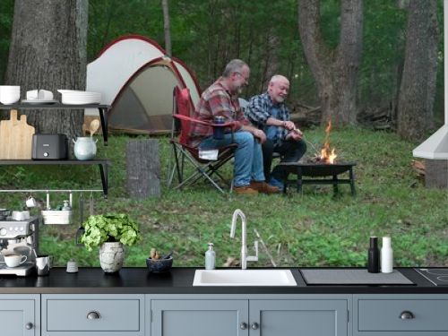 Wide angle view of two gay men building a campfire in a forest talking and laughing with tent in background.